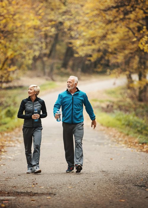 Closeup front view of a senior couple walking in a park after nice jogging workout. They are drinking bottled water and enjoying the view of orange leaves and grass.